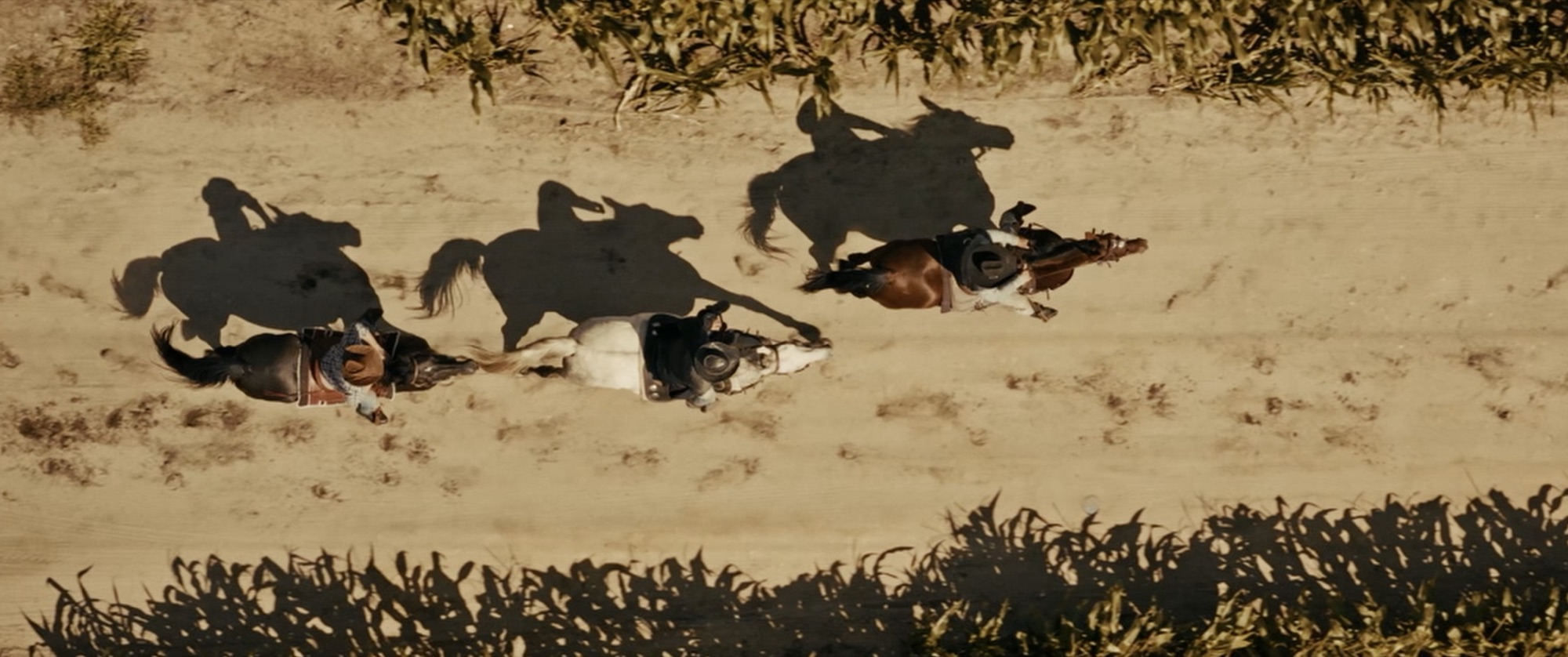 Overhead shot of three horseback riders racing along a sandy track, long shadows across the dust - Amon Amarth